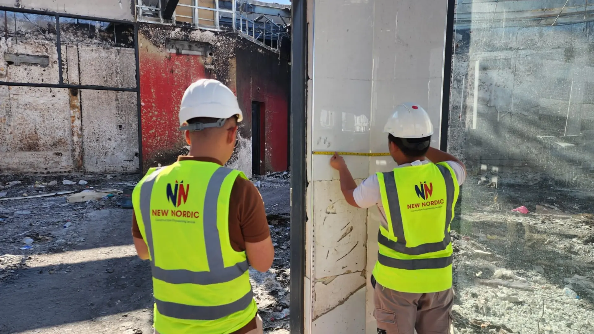 Two New Nordic engineers measuring a fire-damaged wall with a tape measure at a burnt building exterior
